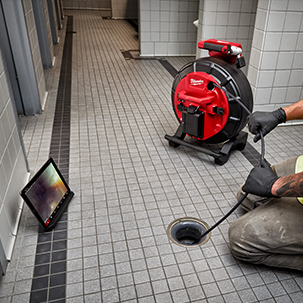 Man feeds cable from Pipeline Inspection System down cleanout in commercial restroom with live video on mobile device nearby.